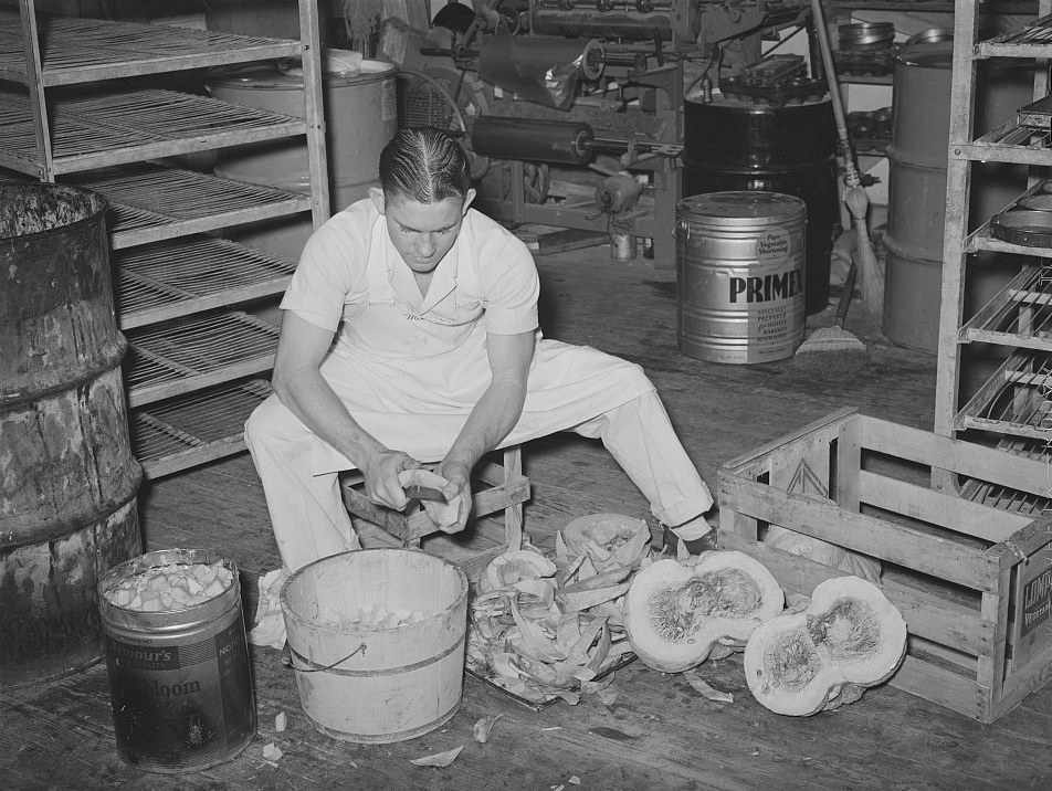 A baker in white overalls cutting up a pumpkin for pies on a stool in a bakery back room, with pumpkin halves and a bucket of peeled pieces, 1939 photograph