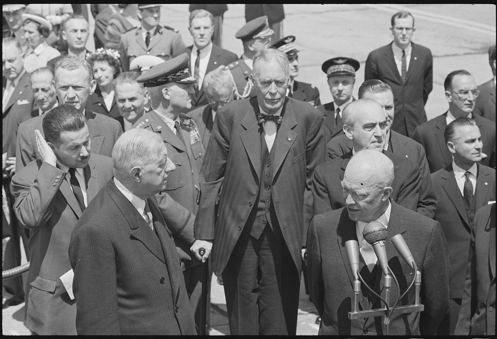 Photograph of Charles de Gaulle in a dark suit standing beside Dwight Eisenhower at a microphone on an airport apron, surrounded by officials, in April 1960