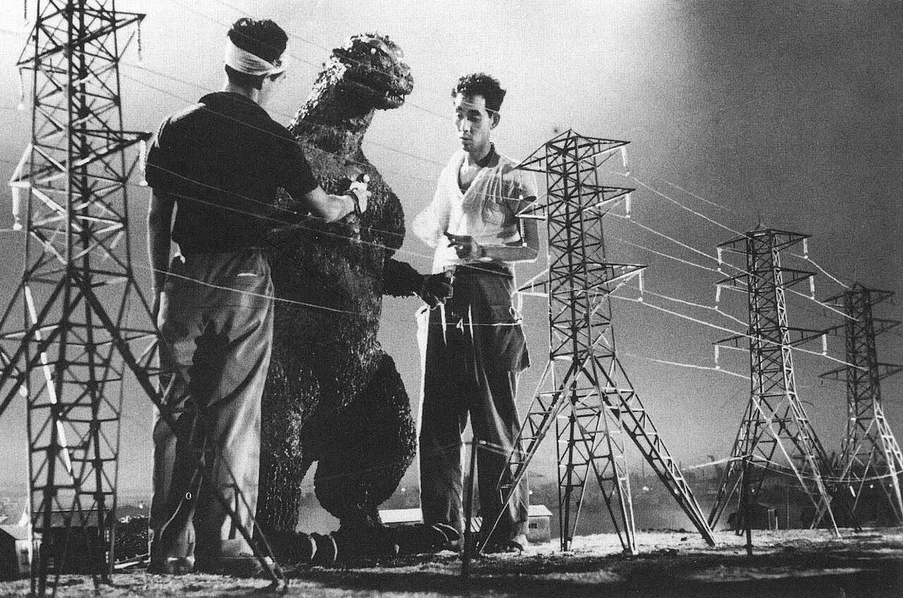 Behind-the-scenes photograph from the making of Godzilla in 1954: two crew members adjust the monster suit on a miniature set of electricity pylons