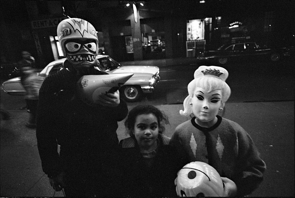 Black-and-white night photograph of three children on a New York street at Halloween in 1964, two of them in moulded plastic masks, one a grinning space monster and one a blonde woman's face