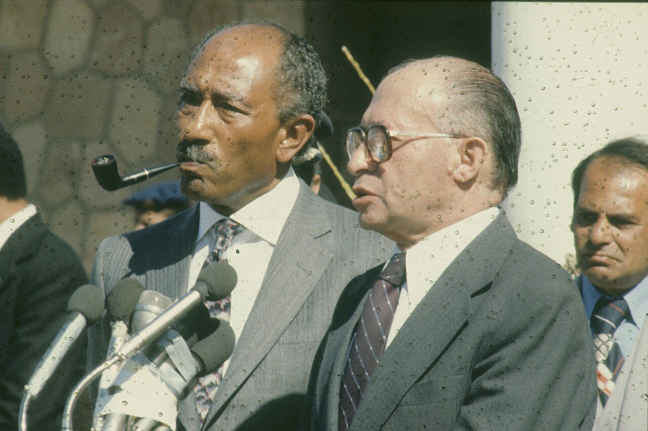 Photograph of Anwar Sadat, pipe in mouth, and Menachem Begin standing at a bank of microphones in the late 1970s