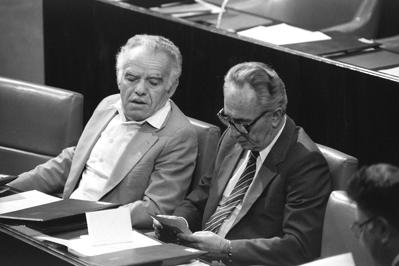 Black-and-white photograph of Yitzhak Shamir and Shimon Peres sitting side by side in the Knesset in the 1980s, Peres reading a note