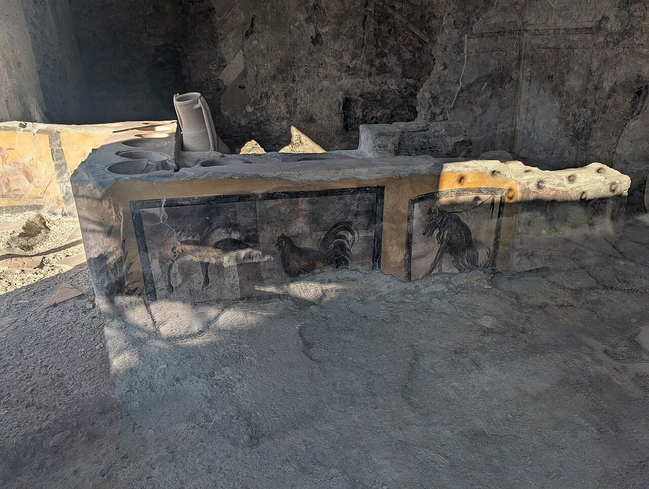 Photograph of the painted food counter in Regio V at Pompeii, with jar openings set into the top and frescoes of hanging ducks, a rooster and a dog on a leash