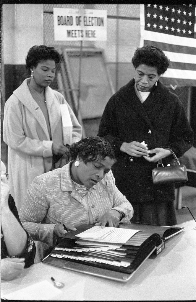 Photograph of three women at a polling place in 1957, one checking a book of registered voters, with a voting booth and an American flag behind them