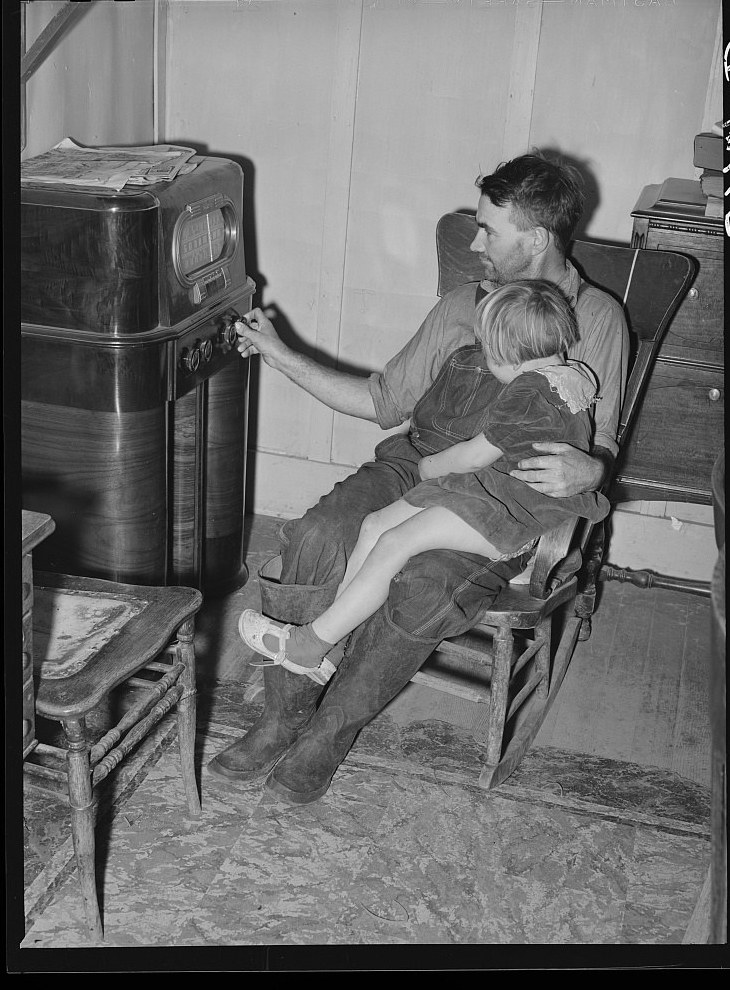 Black and white photograph of a farmer tuning a large wooden console radio with his young daughter on his lap, California, 1940