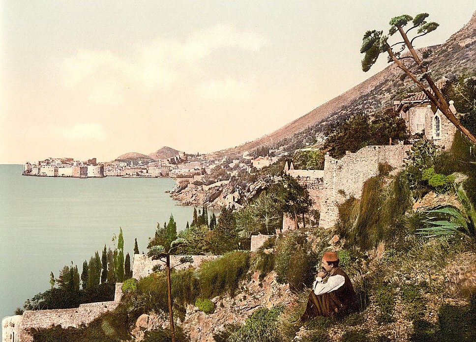 Colour photochrom print of Ragusa (Dubrovnik) seen from the coast road, with the walled old town on its headland and a man sitting on the hillside, about 1890 to 1900